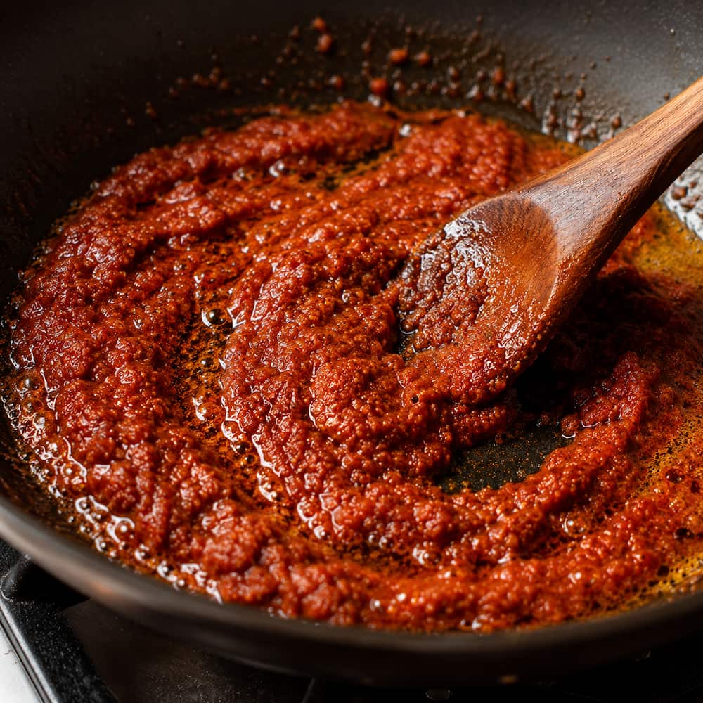 Double-concentrated tomato paste caramelising to deep brick red in a butter and spice base inside a skillet, with oil beginning to separate at the edges